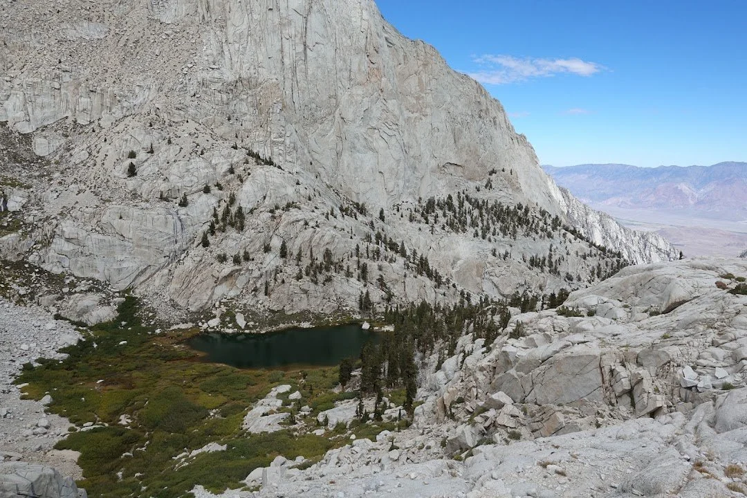 A dark green lake lies below a steep granite wall, edged by conifers and patches of low vegetation.