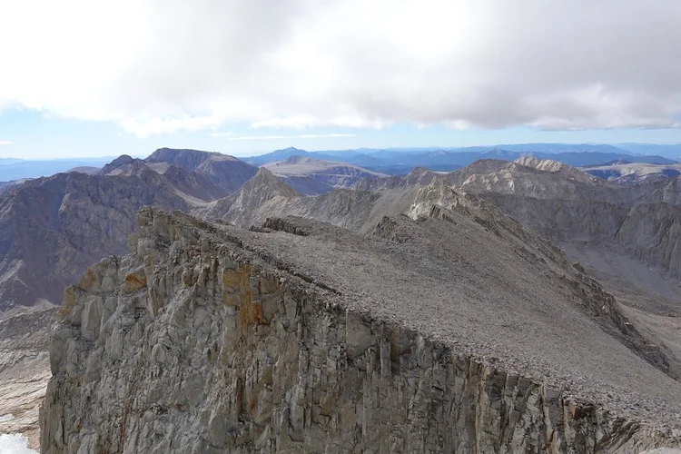A broad, rubble-covered plateau ends in sheer cliffs, with successive mountain ridges beneath a low bank of cloud.