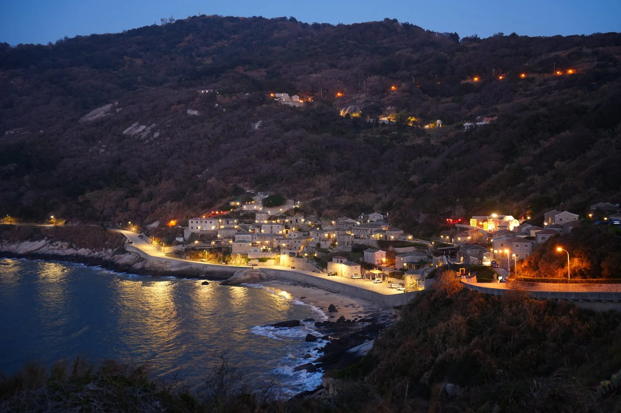 Lights trace the waterfront beneath Qinbi's hillside stone houses at dusk.