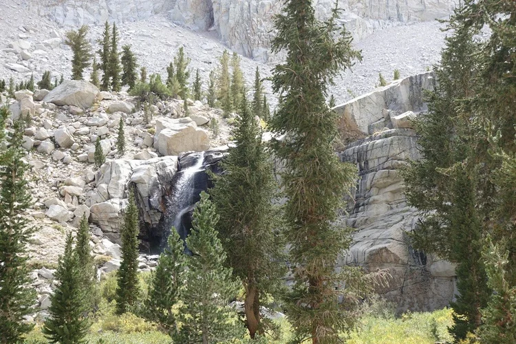 A waterfall slips over dark granite ledges among conifers and scattered boulders.