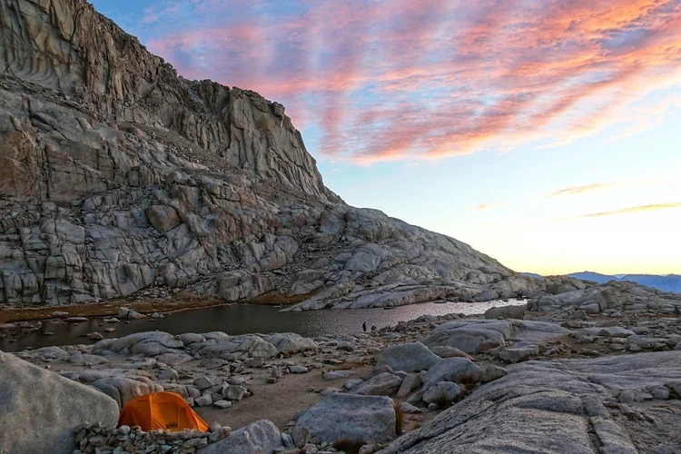 An orange tent stands among granite boulders beside an alpine lake under a sky streaked with pink.