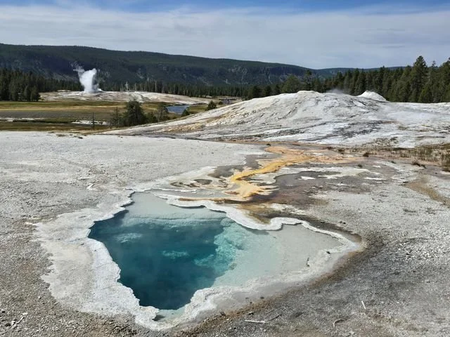 Clear blue water fills Heart Spring, with a geyser plume rising across the basin.