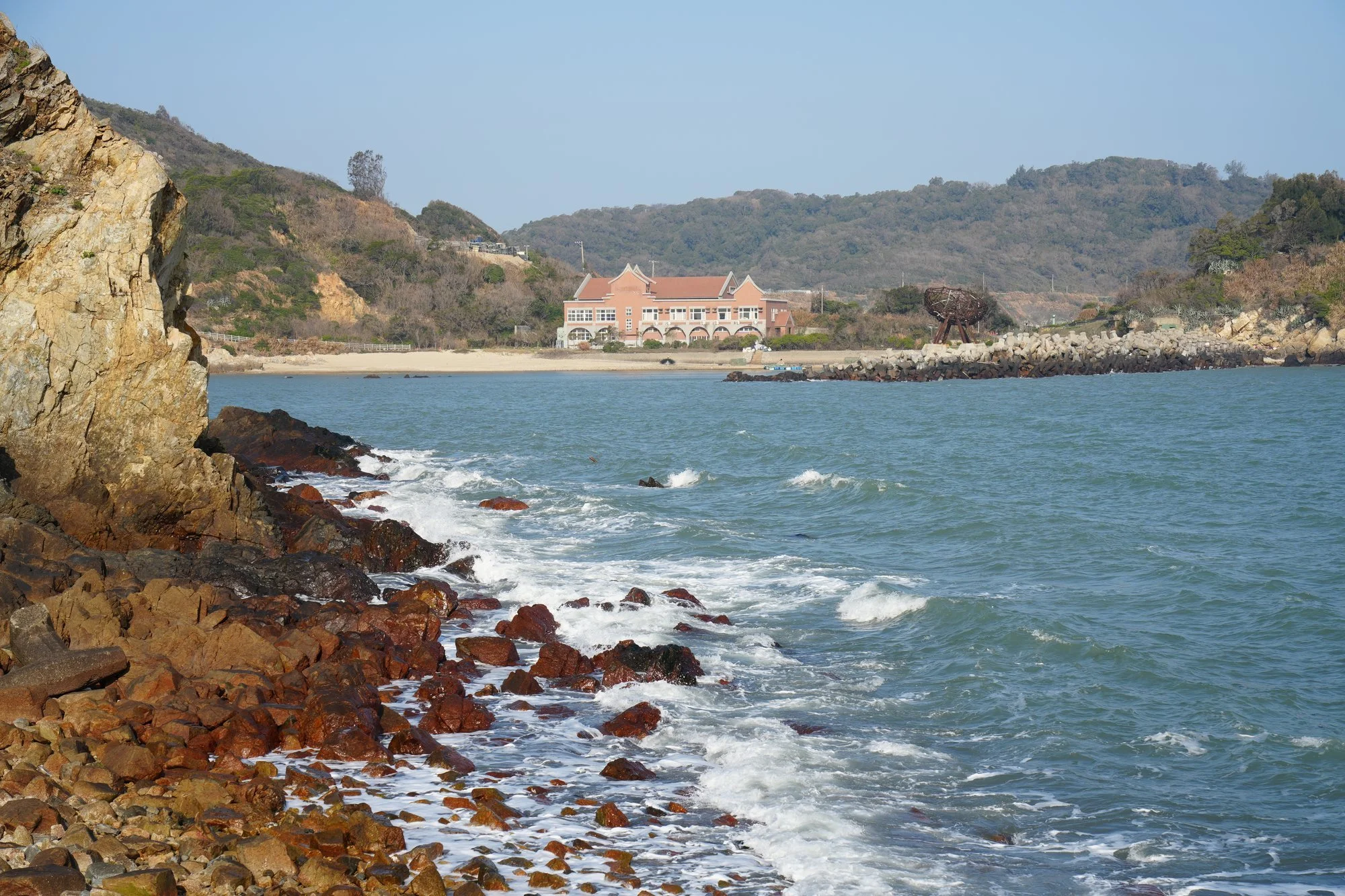 Foam washes over rust-coloured rocks, with a pink building across the bay on Nangan.