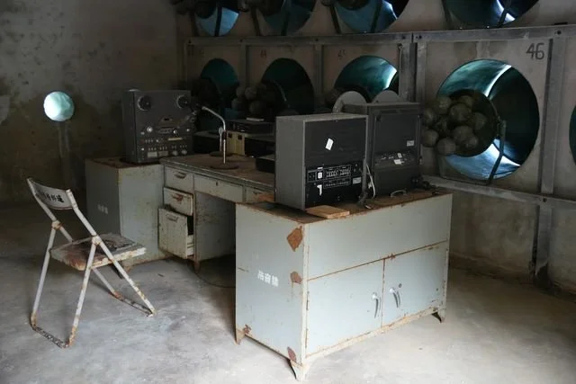 Recording equipment stands beneath rows of loudspeakers in a weathered room on Beigan.