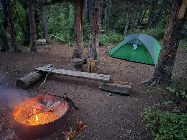 A fire glows beside a green tent and a rough log bench in a forest clearing.