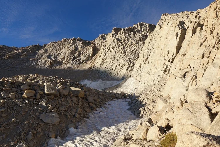 Compacted snow fills a narrow passage between granite boulders beneath a sunlit cliff.