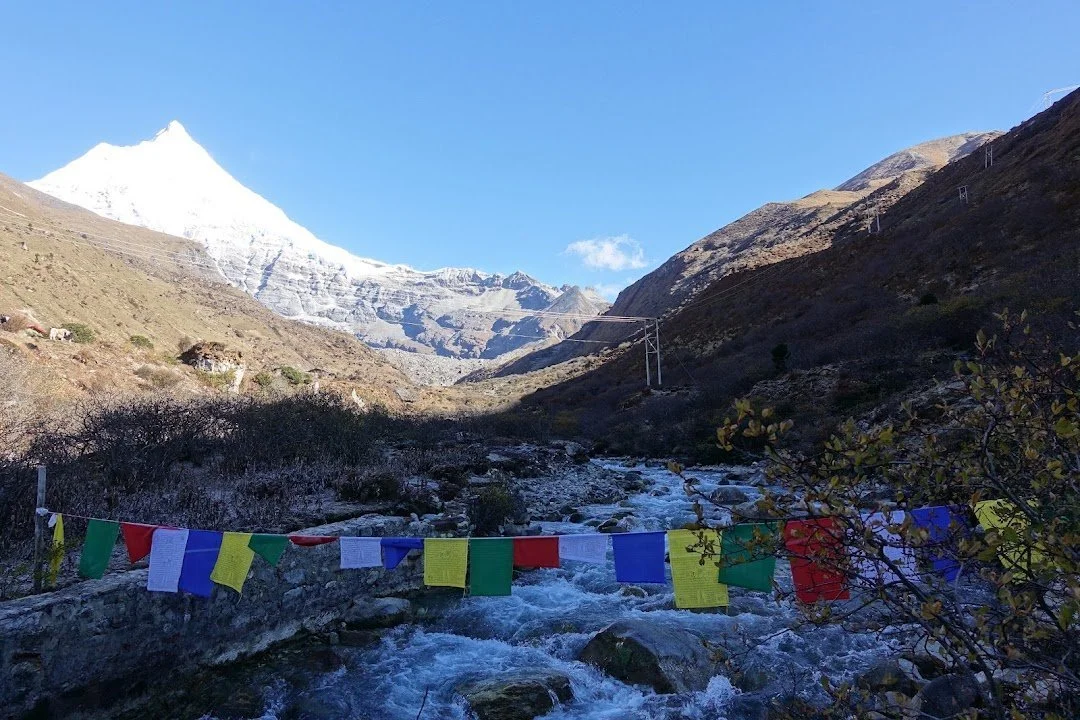 Prayer flags span the rushing stream, with a white peak rising at the head of the valley.