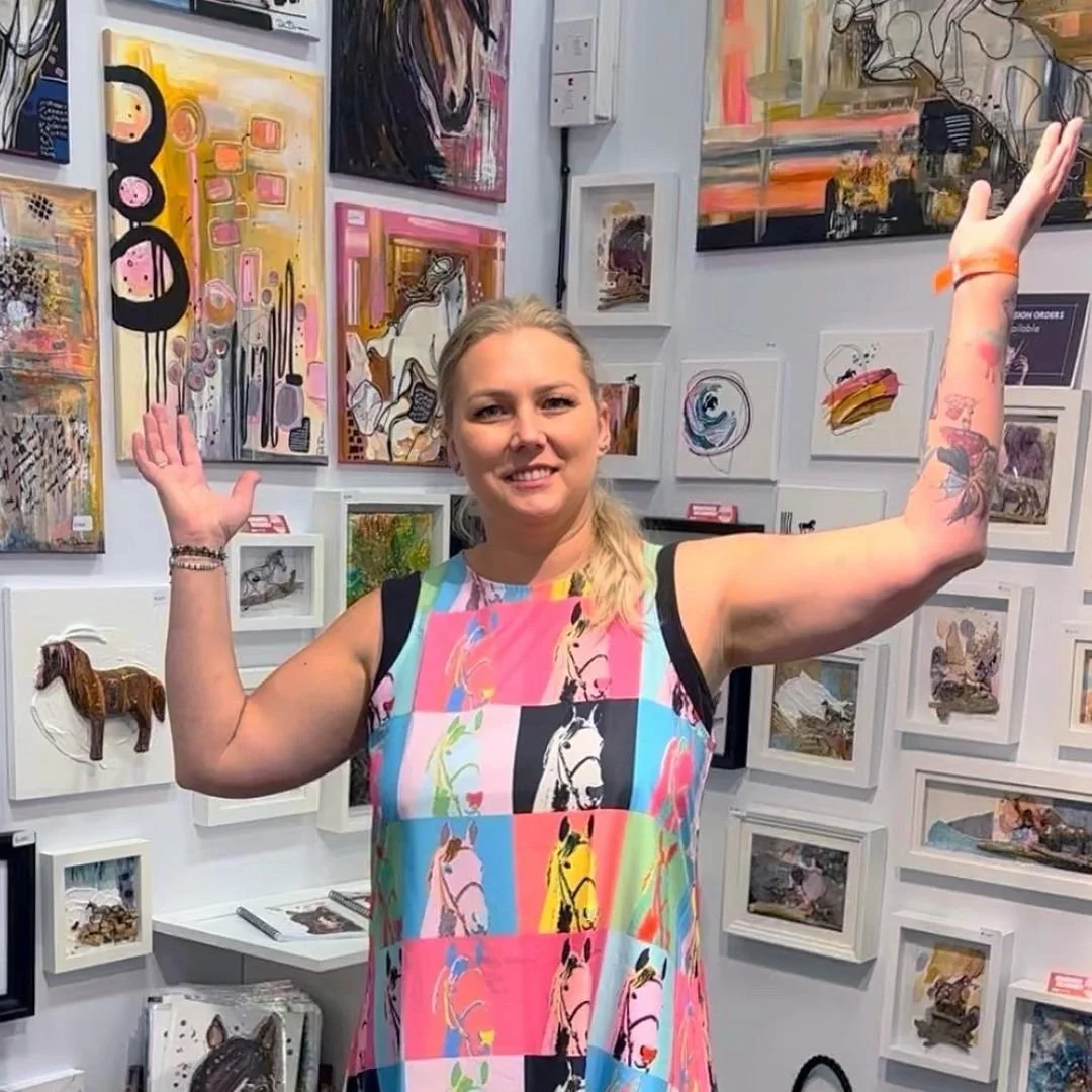 Karolina Di Duca with her arms raised in her gallery, surrounded by her framed work