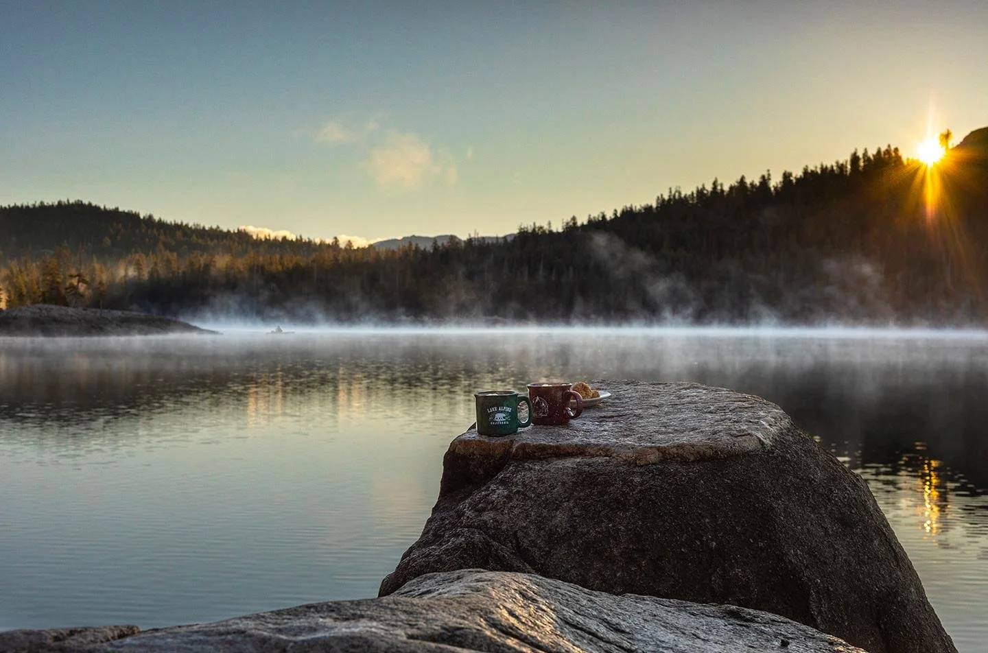 Two enamel camp mugs, one green and one red, on a granite boulder above a still lake at sunrise, with mist on the water, a forested ridge behind and a lone boat in the distance.
