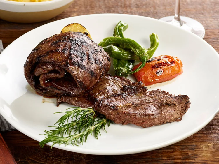 Entraña skirt steak grilled over open fire, served on a white plate with grilled shishito peppers and tomato