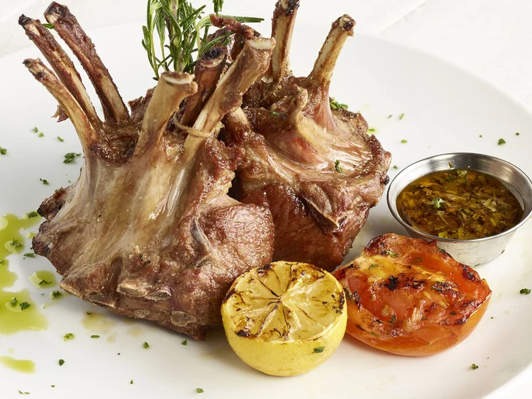 Rack of lamb standing upright on a white plate with grilled lemon, tomato and chimichurri