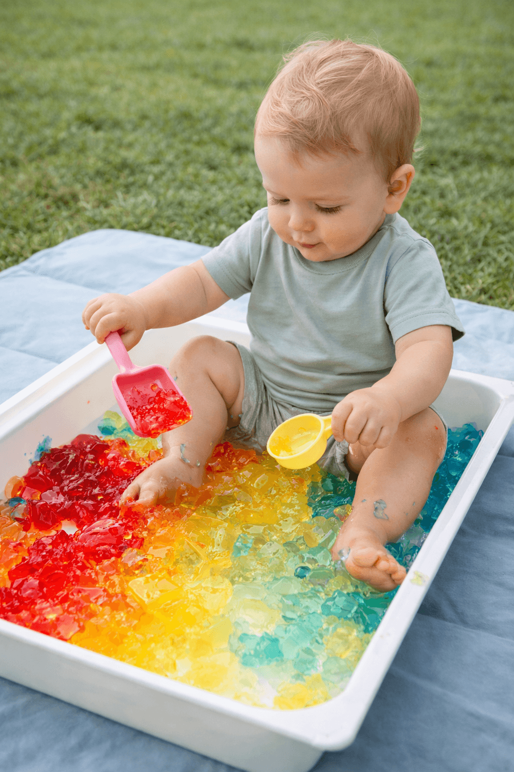 Toddler squishing and scooping colourful jelly in a shallow tray outdoors
