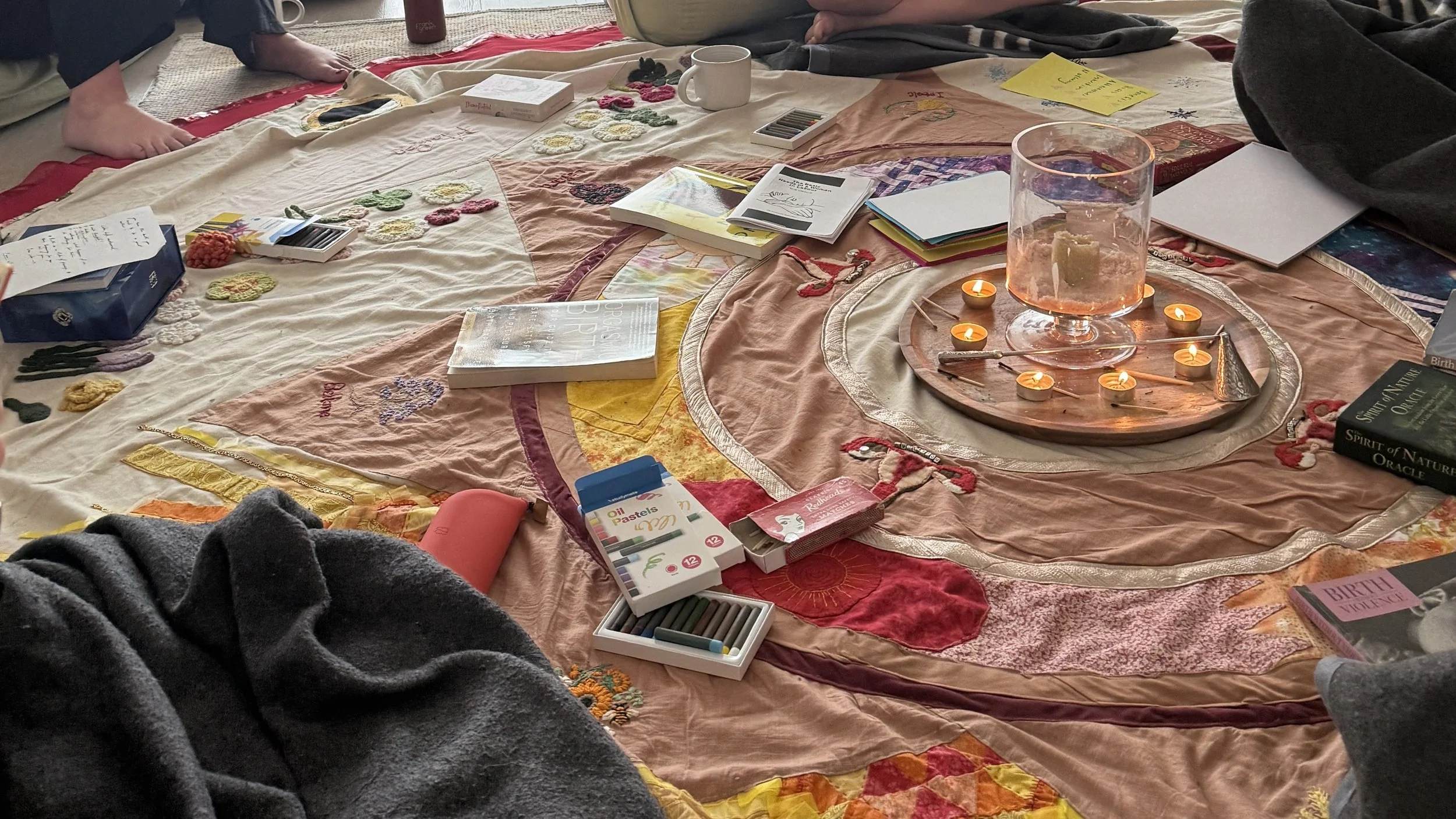 An embroidered cloth laid out on the floor for a women's circle, with candles, oil pastels and open notebooks