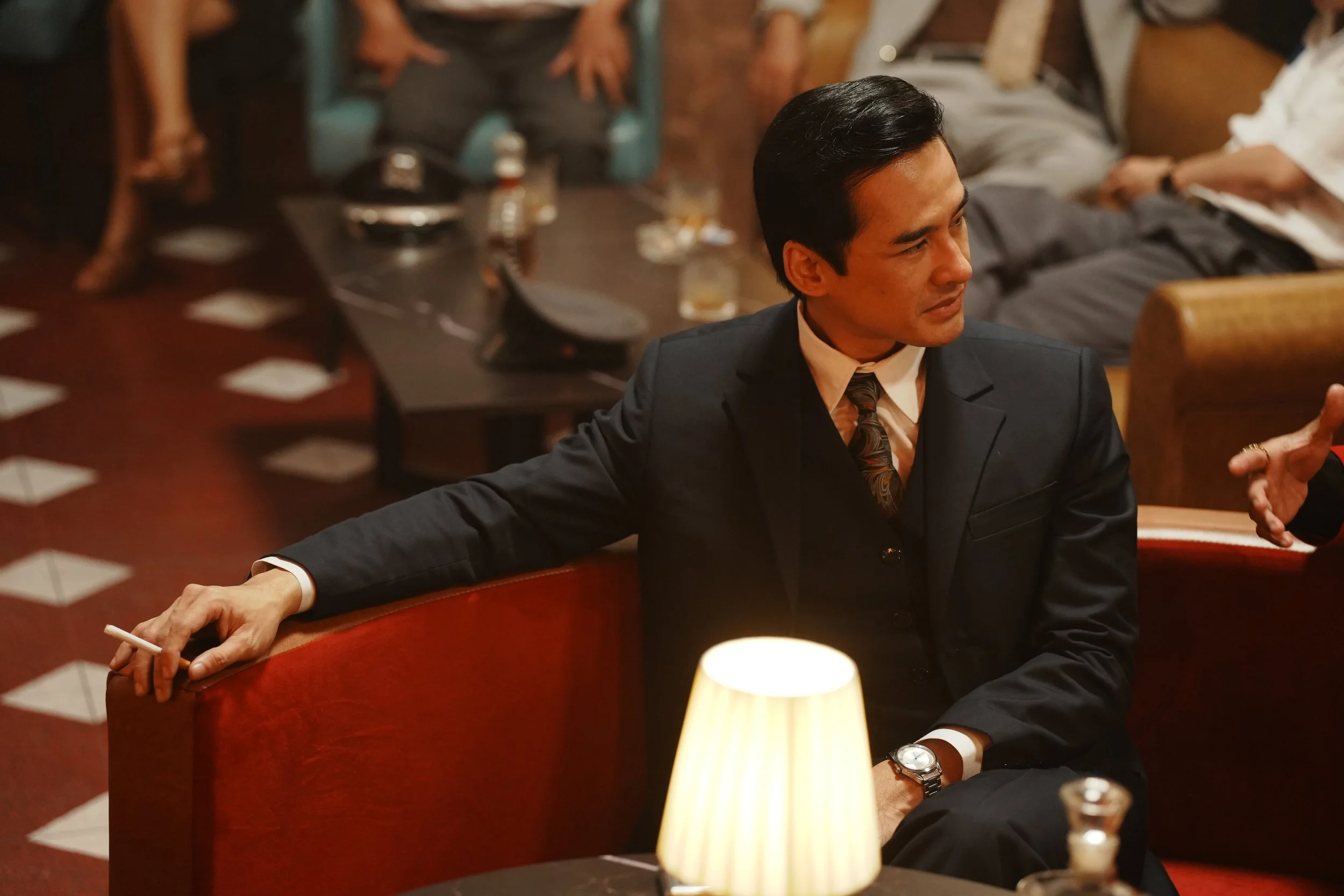 A man in a three-piece suit lounges in a red velvet booth in a 1970s Saigon club, in a still from Madame in Saigon