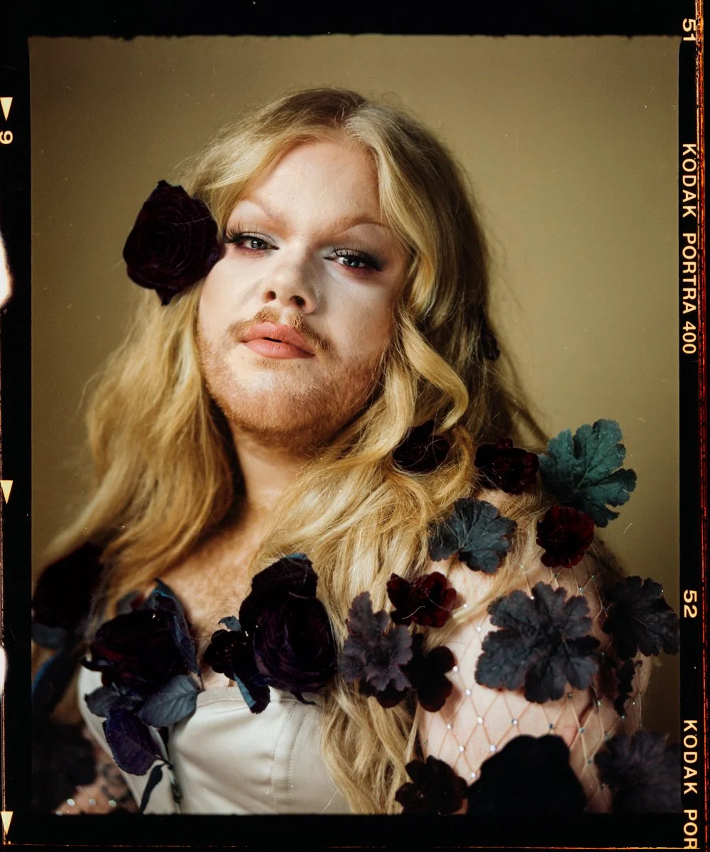 Drag portrait: Grace Shushh facing camera in a dark floral collar under warm light.