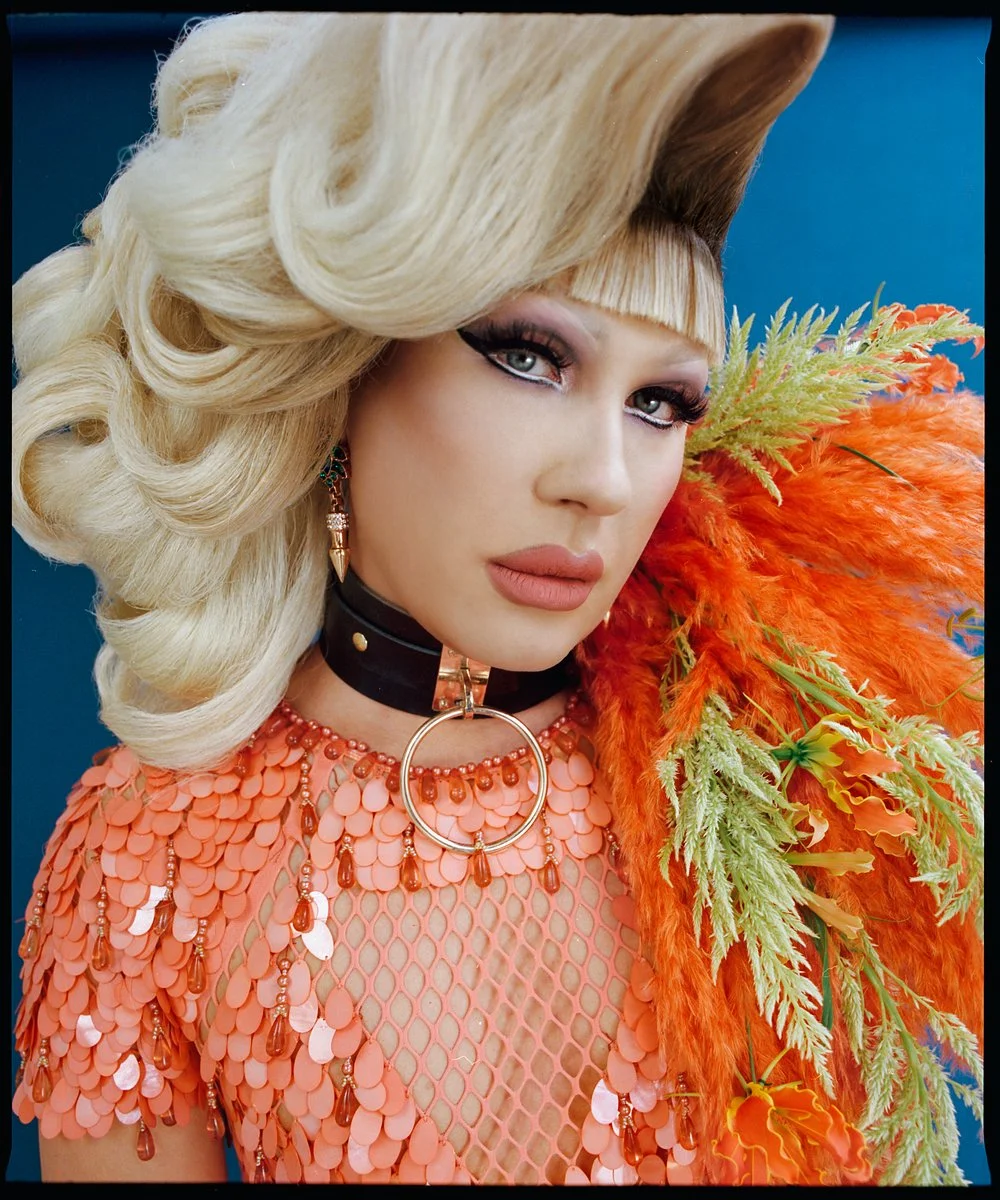 Drag portrait: close-up with a blonde bouffant and orange feathers.