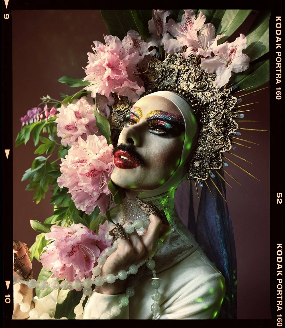 Drag portrait: Virgin X in profile under a pink floral headdress on a deep red backdrop.