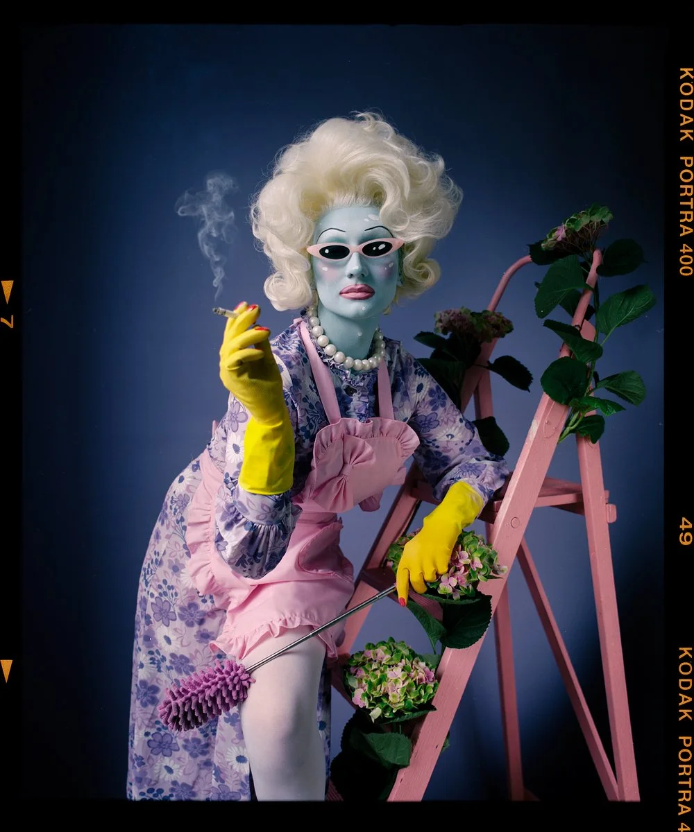 Drag portrait: Juno Birch in pink gloves and a striped apron beside a pink stepladder.
