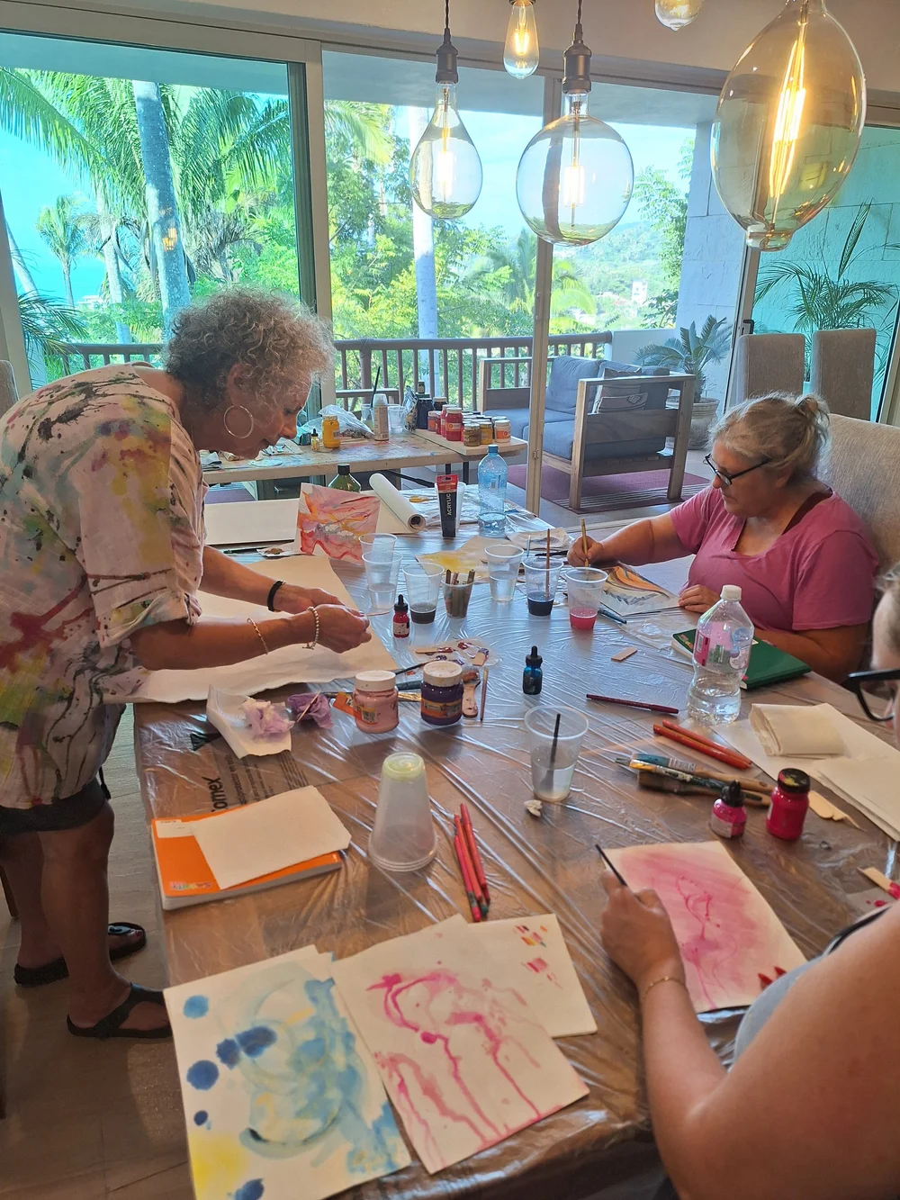 Guests painting at a long table overlooking palms and the ocean