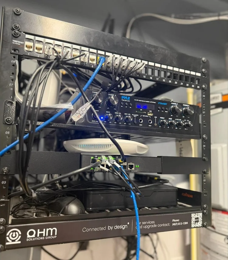 Café equipment rack with patch panel, network switch and audio amplifier labeled by Ohm Solutions Group