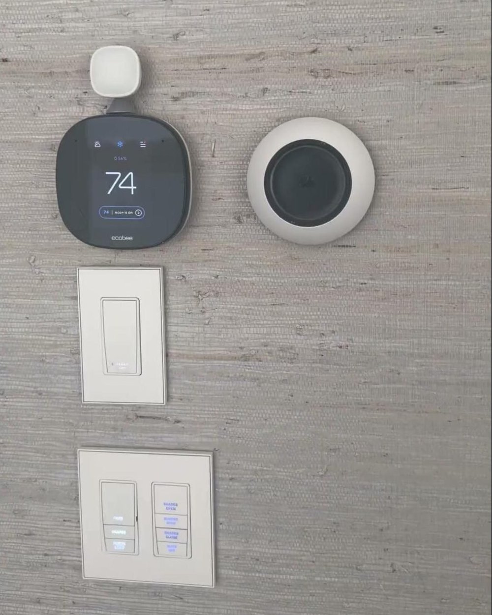 Smart thermostat with remote room sensor above engraved, backlit lighting and shade keypads on a grasscloth wall