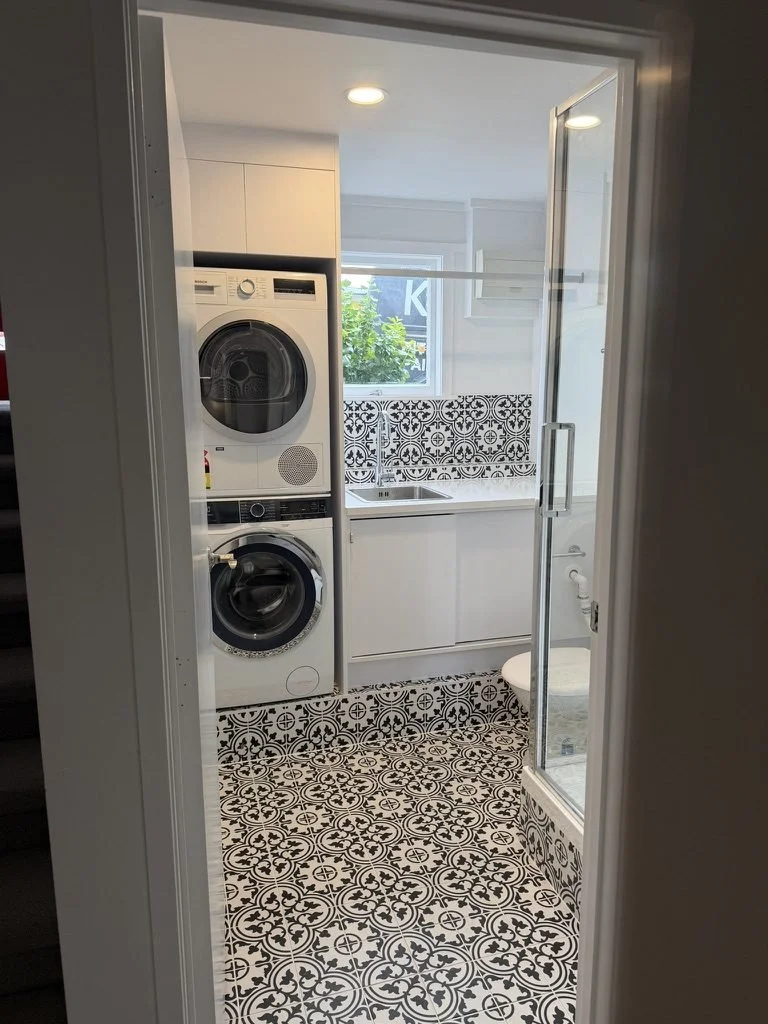 Finished Belmont laundry bathroom seen through the glass shower door, stacked machines and encaustic floor