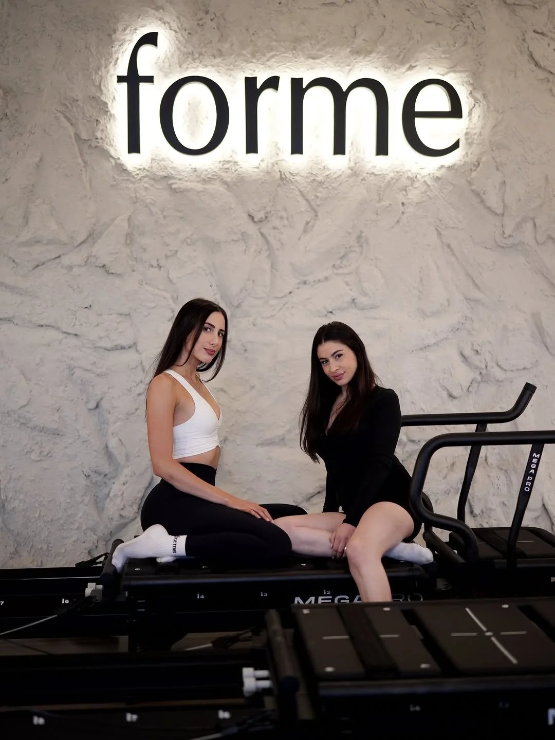 Two people sitting on a Lagree machine in front of the lit forme wall at Forme Burbank