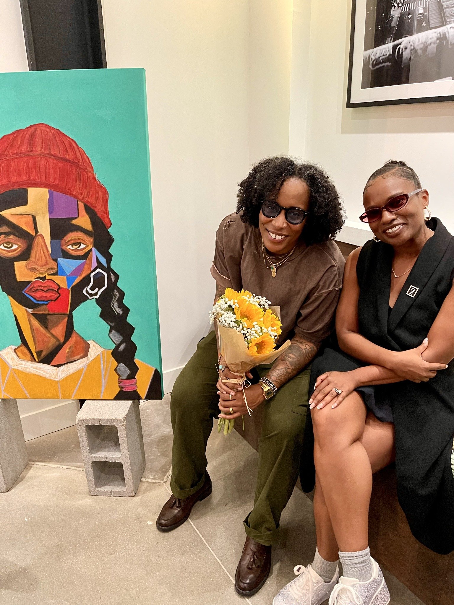 Wyntress X’ion and Allison Retina Stewart-Creeks seated beside a turquoise portrait at In Every Form.