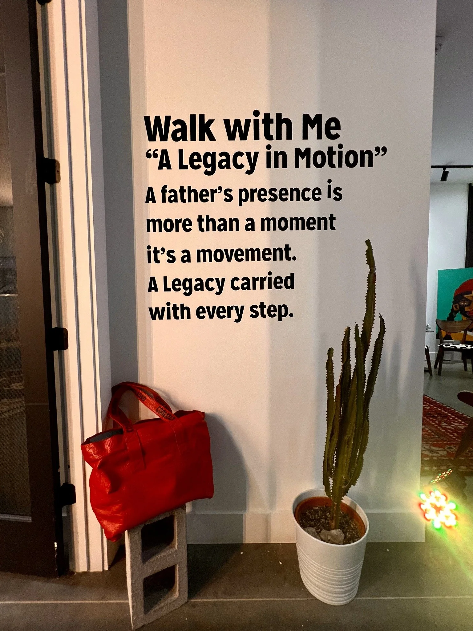 Walk With Me — A Legacy in Motion installation, with a red bag and a potted plant beneath the wall text.