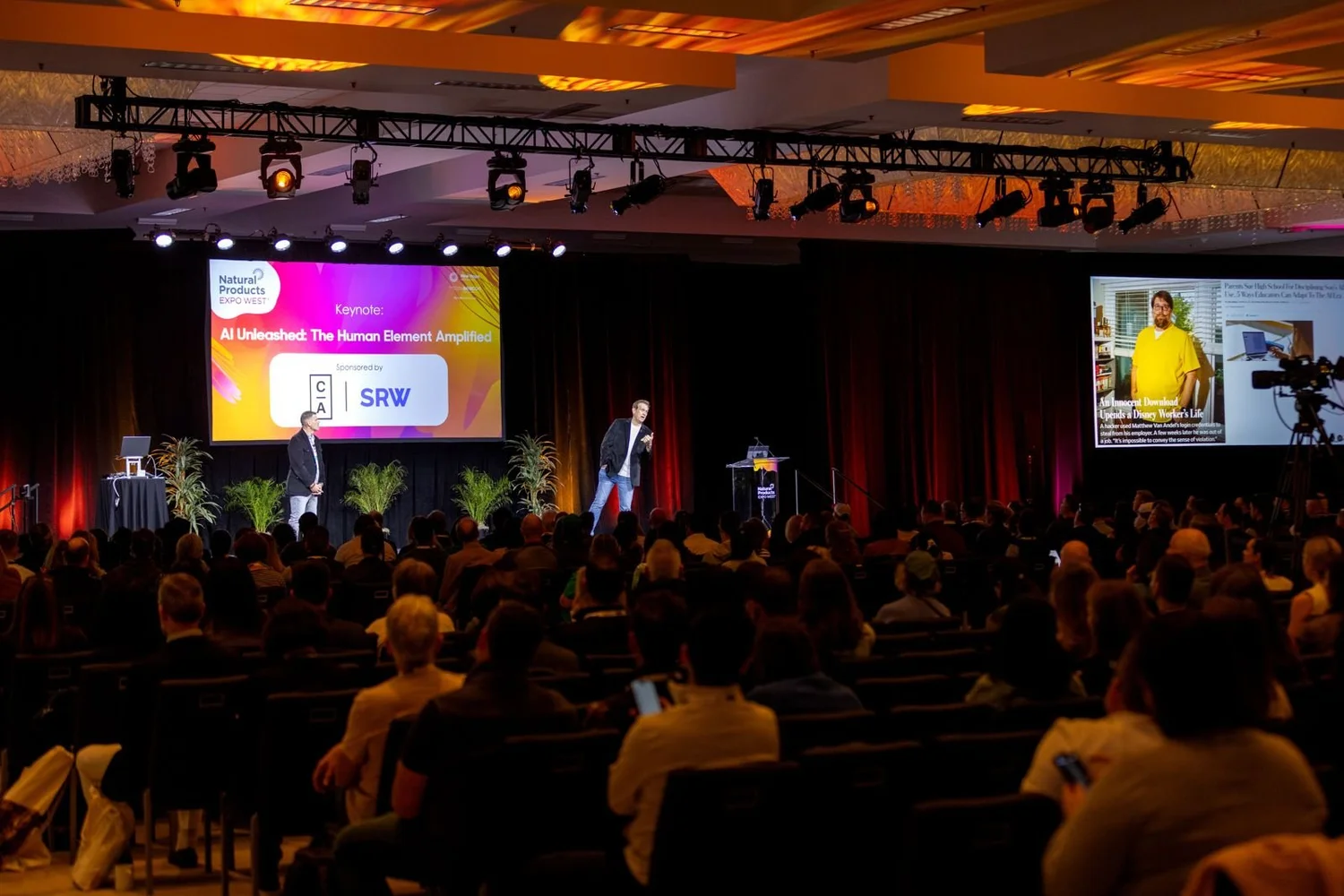 Wide view of a packed ballroom audience facing a stage and large screens