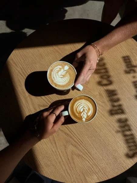 Two Sunrise Brew lattes held over a sunlit table