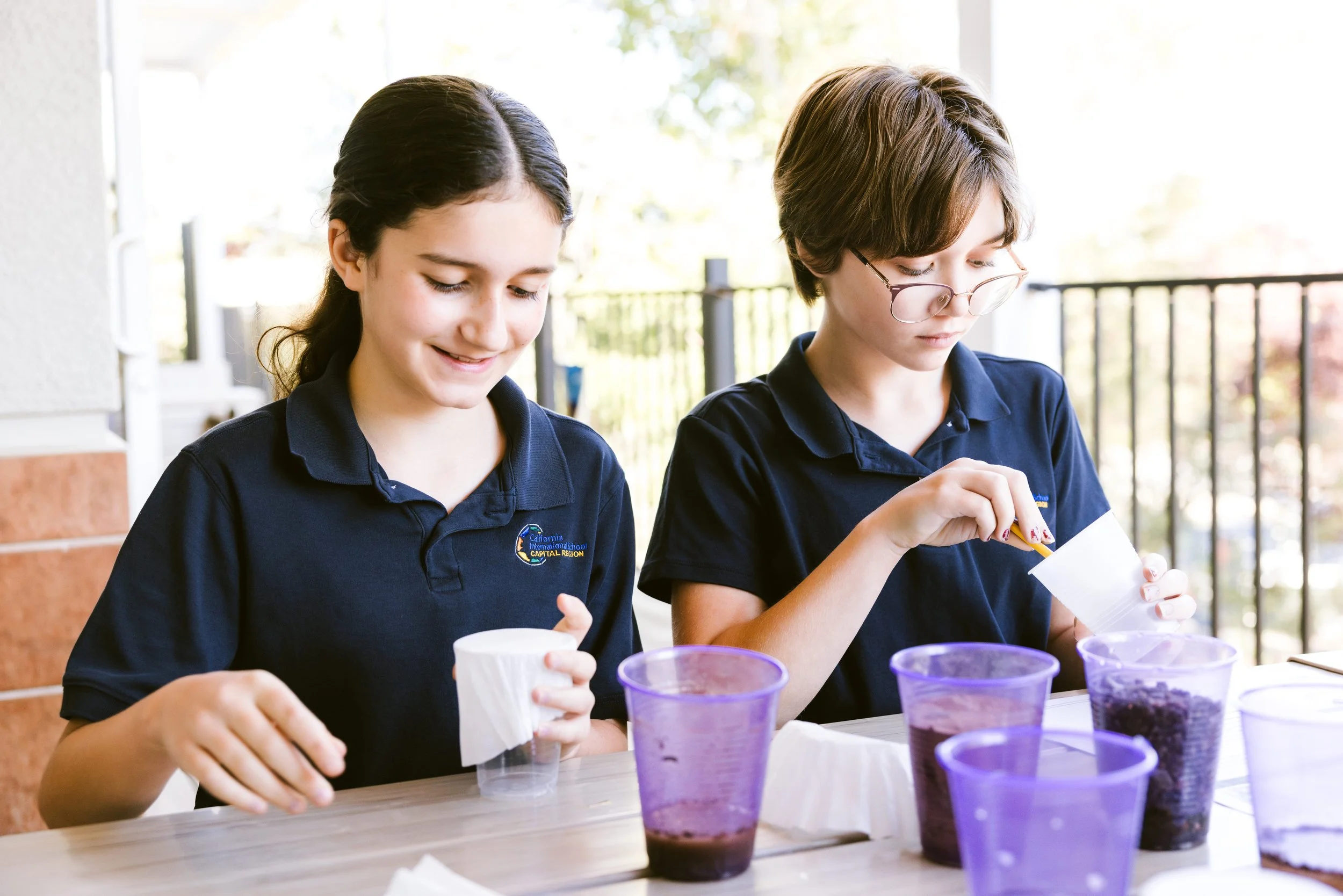Two students in navy school uniforms conducting hands-on science experiment with beakers and purple liquid