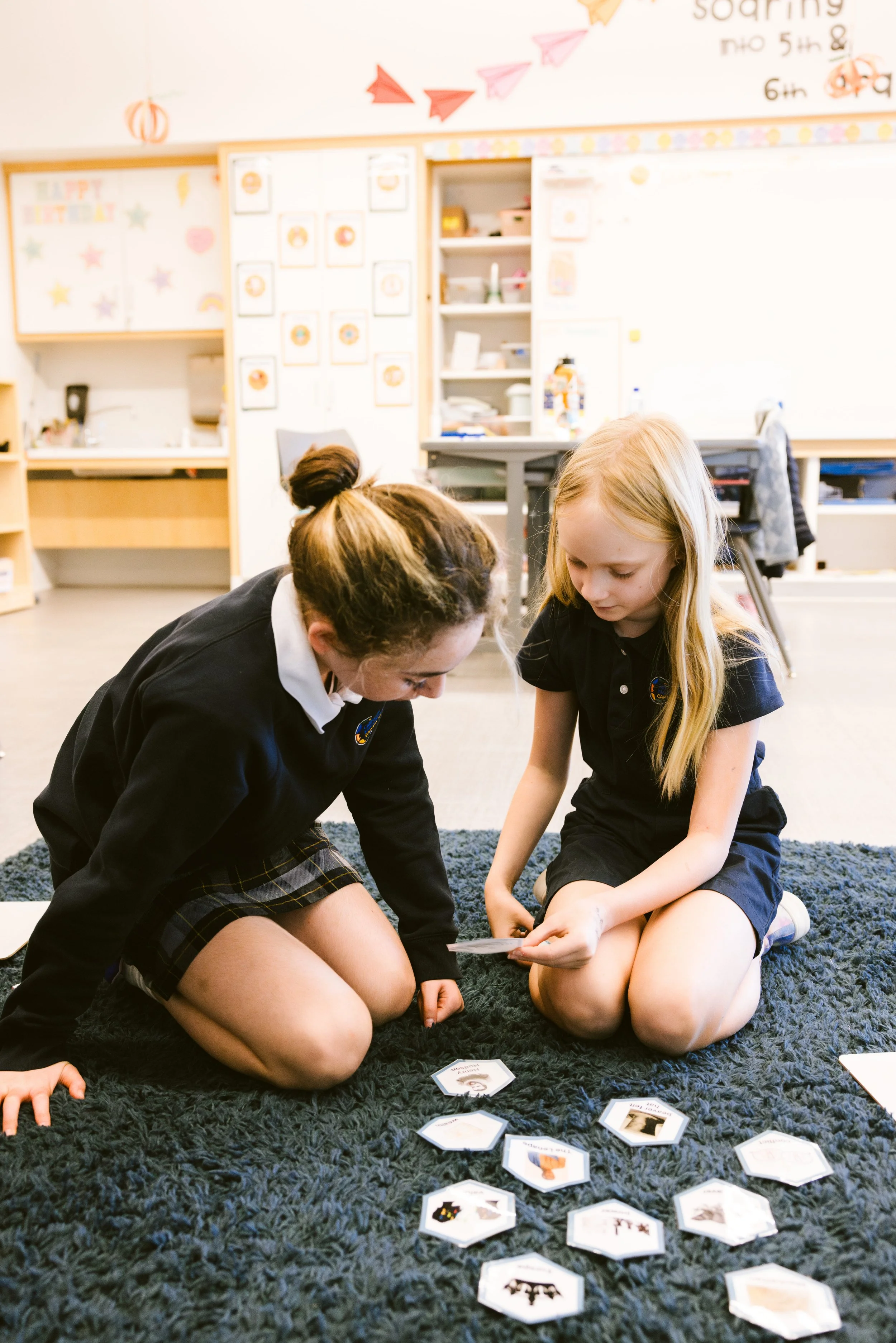 Two female students in school uniforms reading together on carpet in Montessori classroom with shelves in background