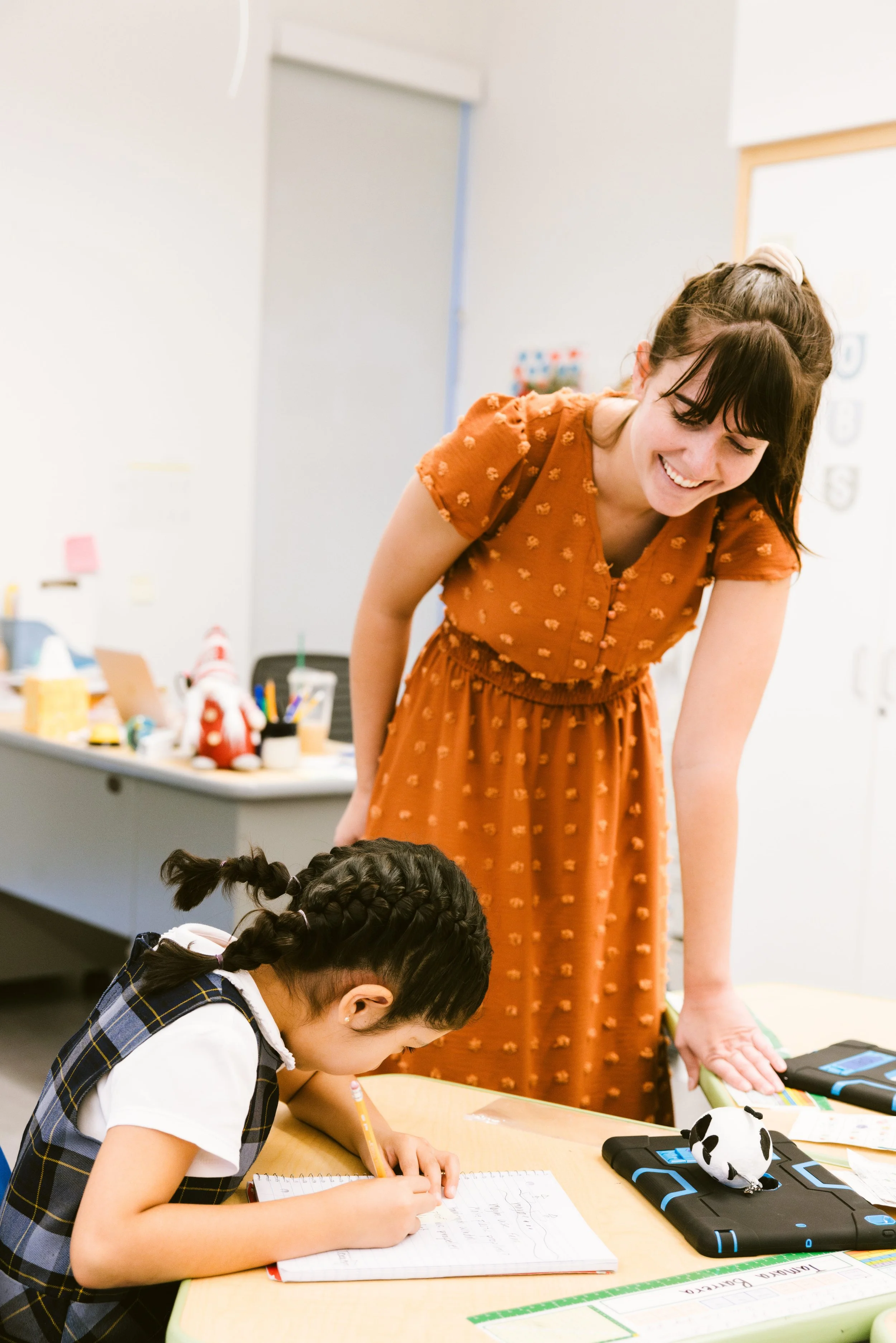 Teacher in orange polka dot dress working one-on-one with young student at learning table in bright classroom
