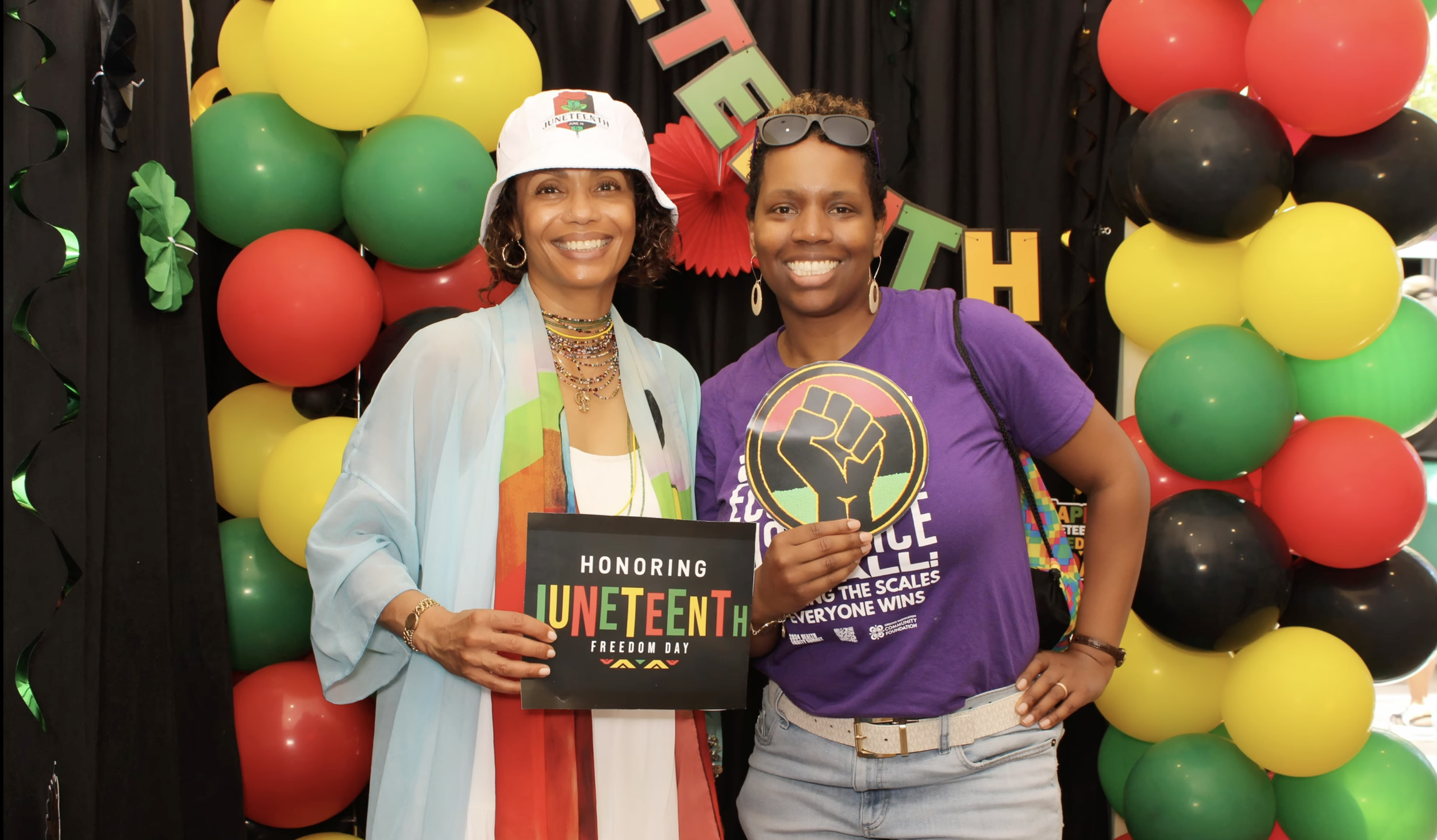 Two middle aged Black women smiling for a photo on Juneteenth. They hold a sign that says “Honoring Juneteenth Freedom Day”. In the background is a black backdrop with red, yellow, and green balloons.