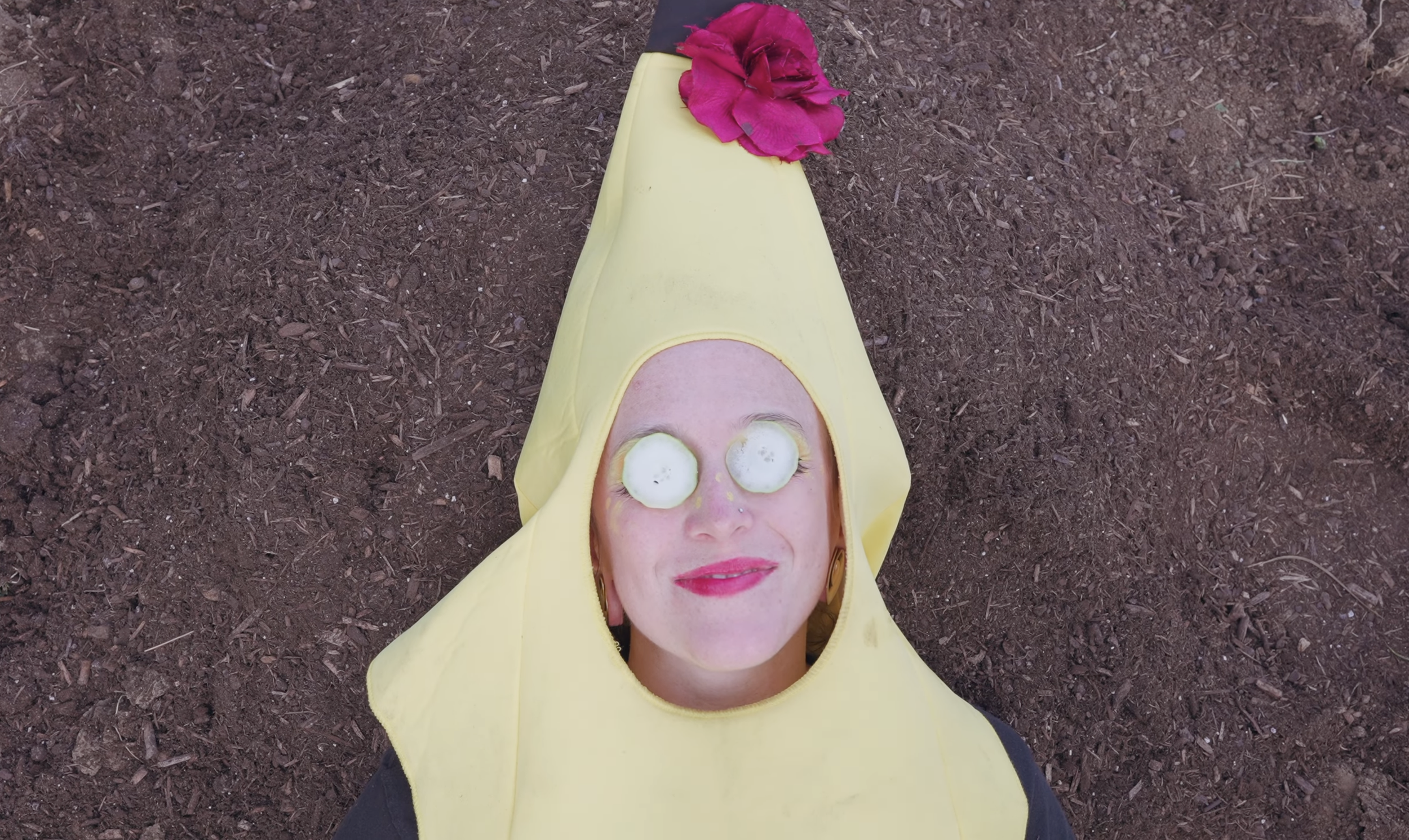 A woman dressed up like a banana laying in the dirt with cucumbers over her eyes and red lipstick on as if she is at the spa.