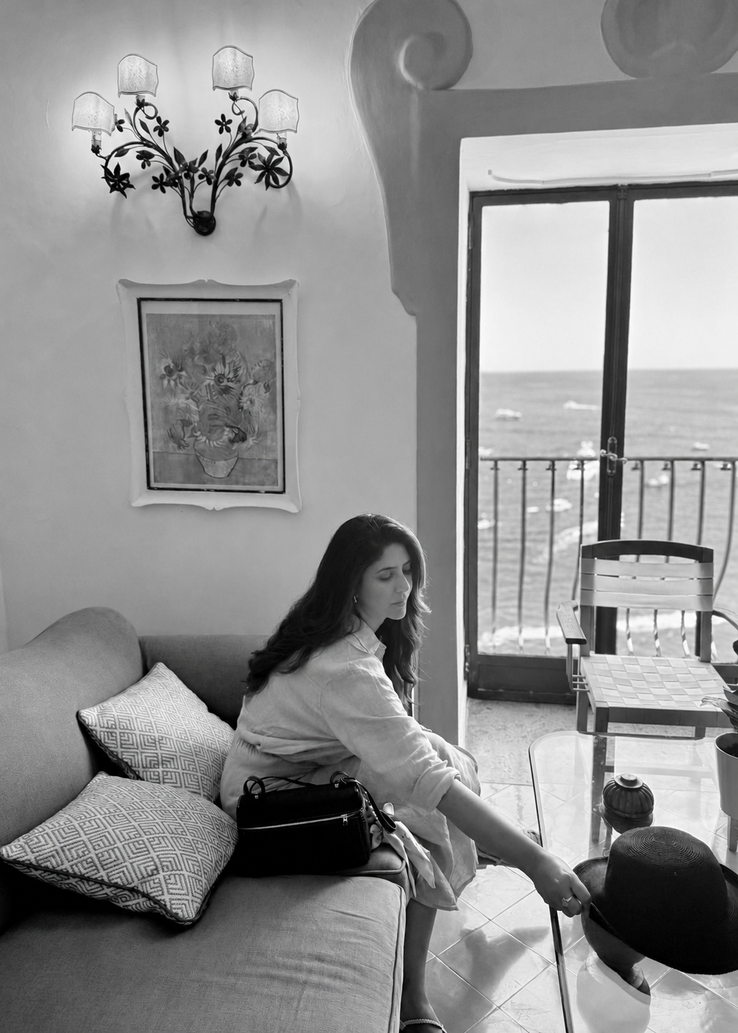 Black-and-white candid portrait of Karishma seated beside a window, reaching towards a table.
