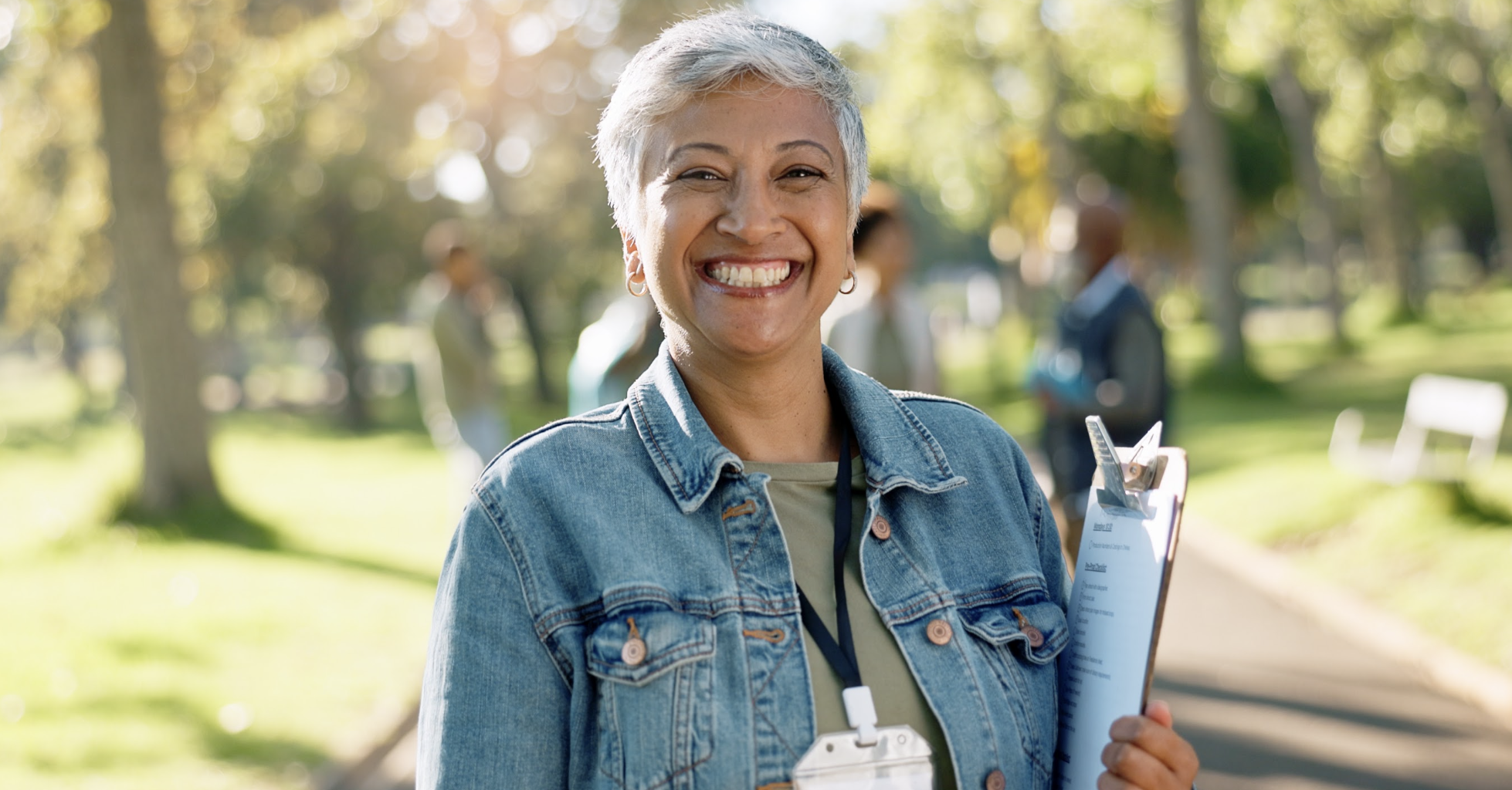 Smiling woman holding a clipboard and wearing a lanyard in a sunny park.