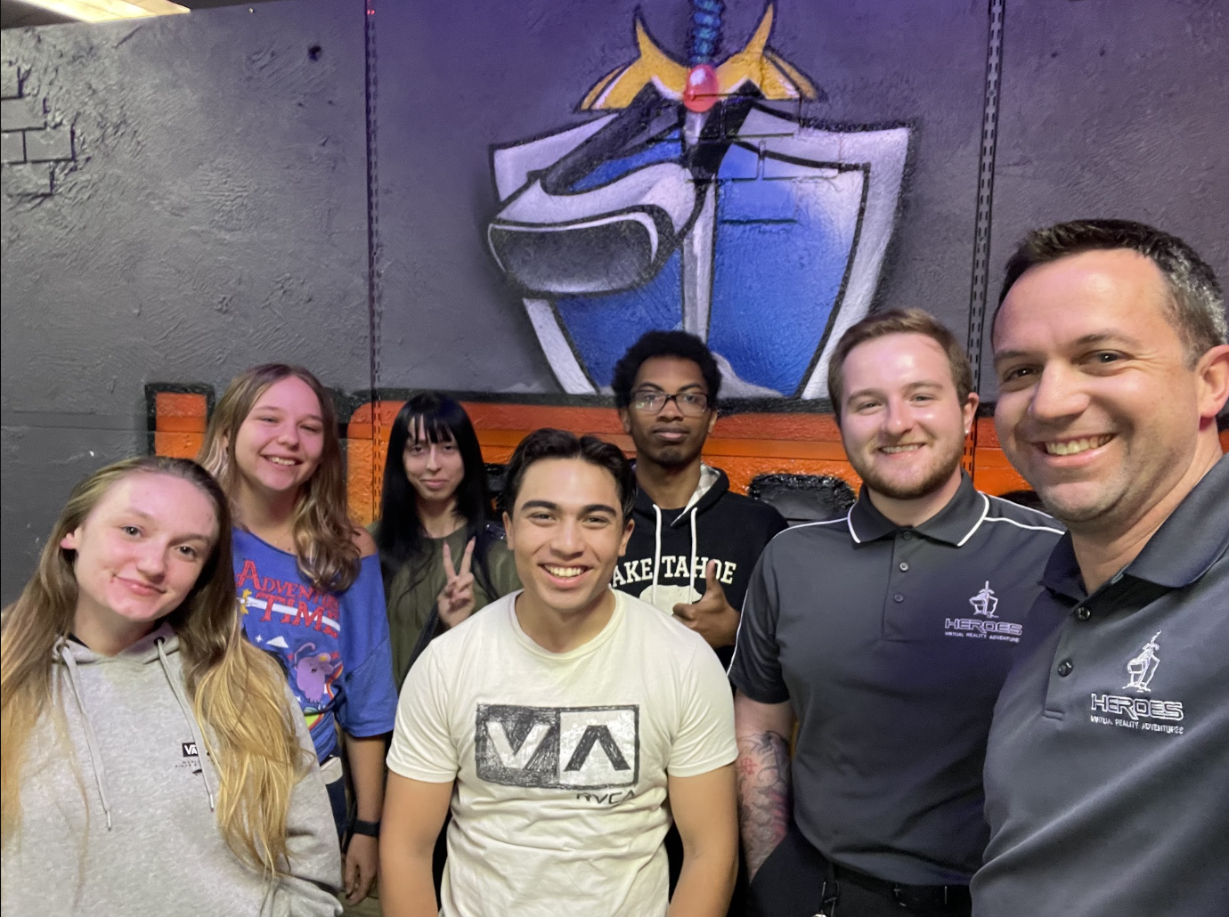 Guests and staff smiling for a group selfie in front of the HEROES VR Adventures wall logo.