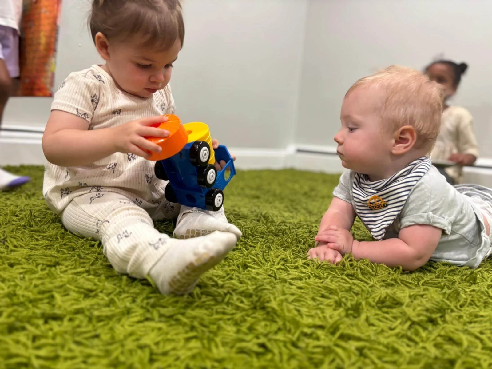 Two infants exploring toys together on a green play mat