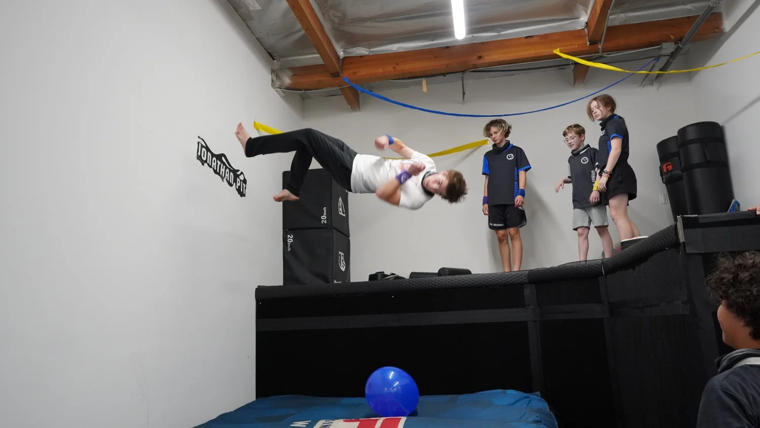 A student practicing an aerial movement above padded equipment with other students nearby