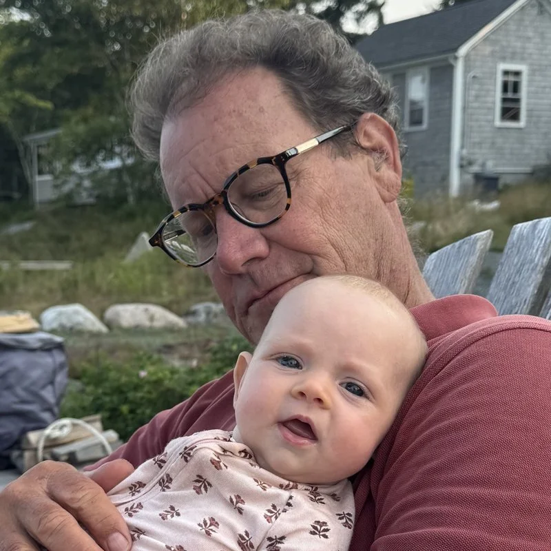 Tom Haslett sitting in an Adirondack chair by the water, holding his grandchild.