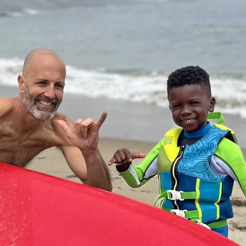 Jeremy Litchfield holding a red surfboard on a foggy Maine beach beside a young boy in a wetsuit.