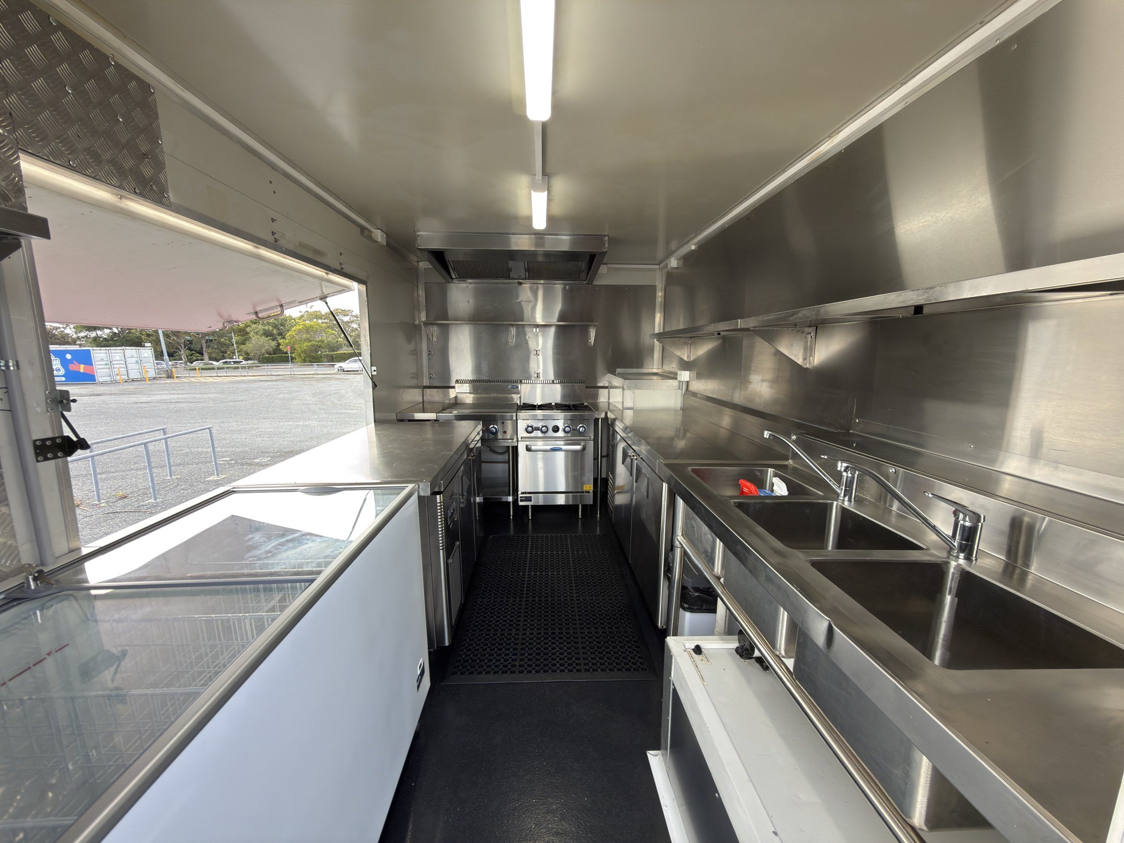 Wide view of the full commercial kitchen with stainless benches, refrigeration, sinks, cooking range, oven, griddle and extraction hood