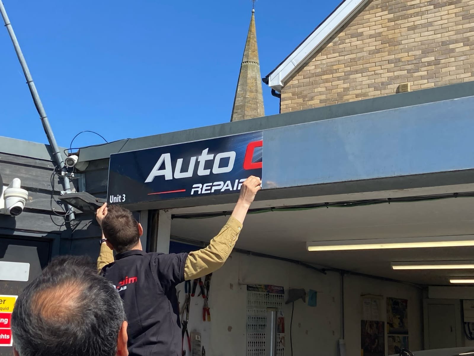 Auto Claim Repair Lab Team Members Lifting Logo Banner on the Garage Front