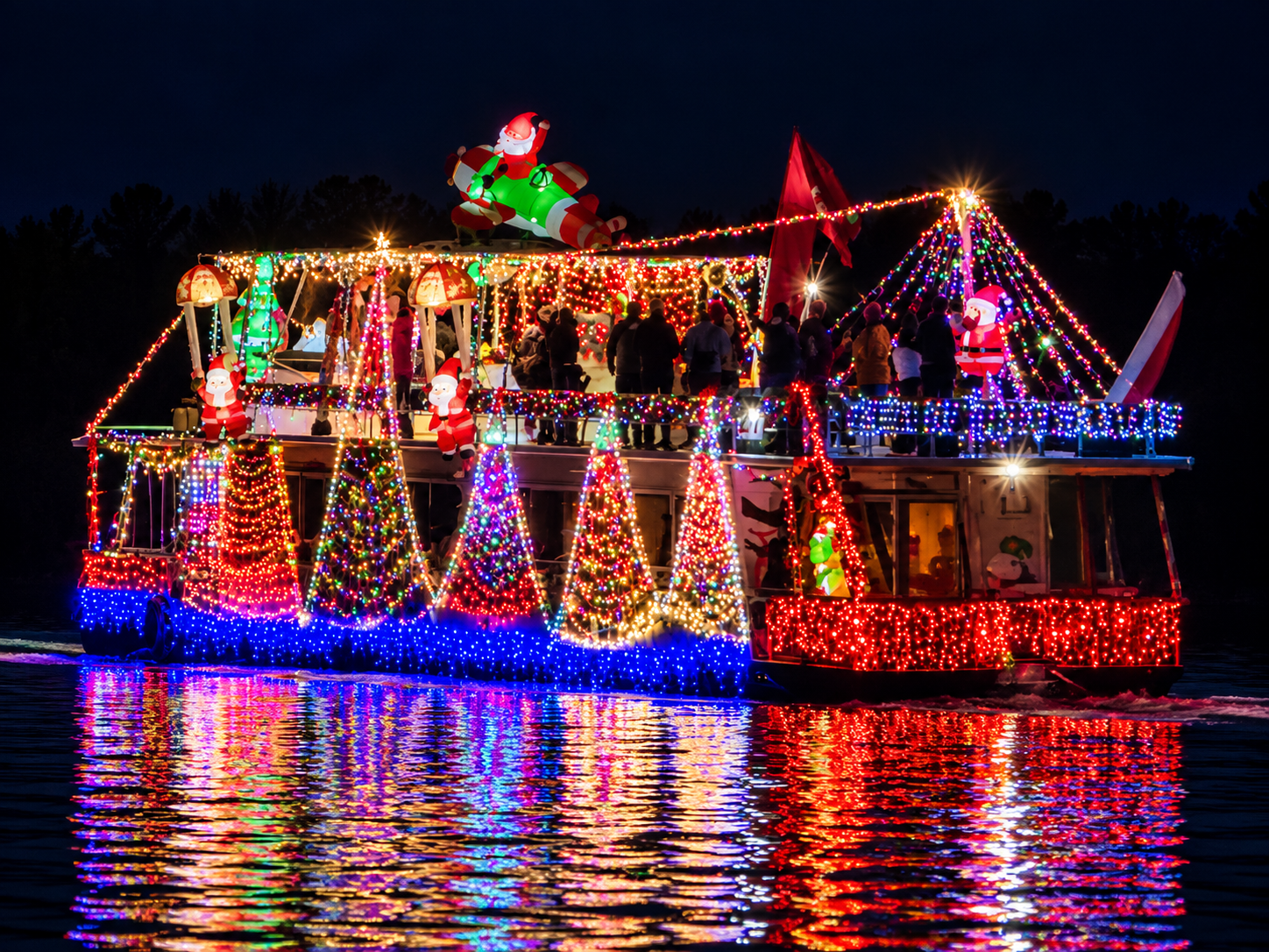 A houseboat covered in colorful Christmas lights and inflatable Santas cruising at night, reflected in the river