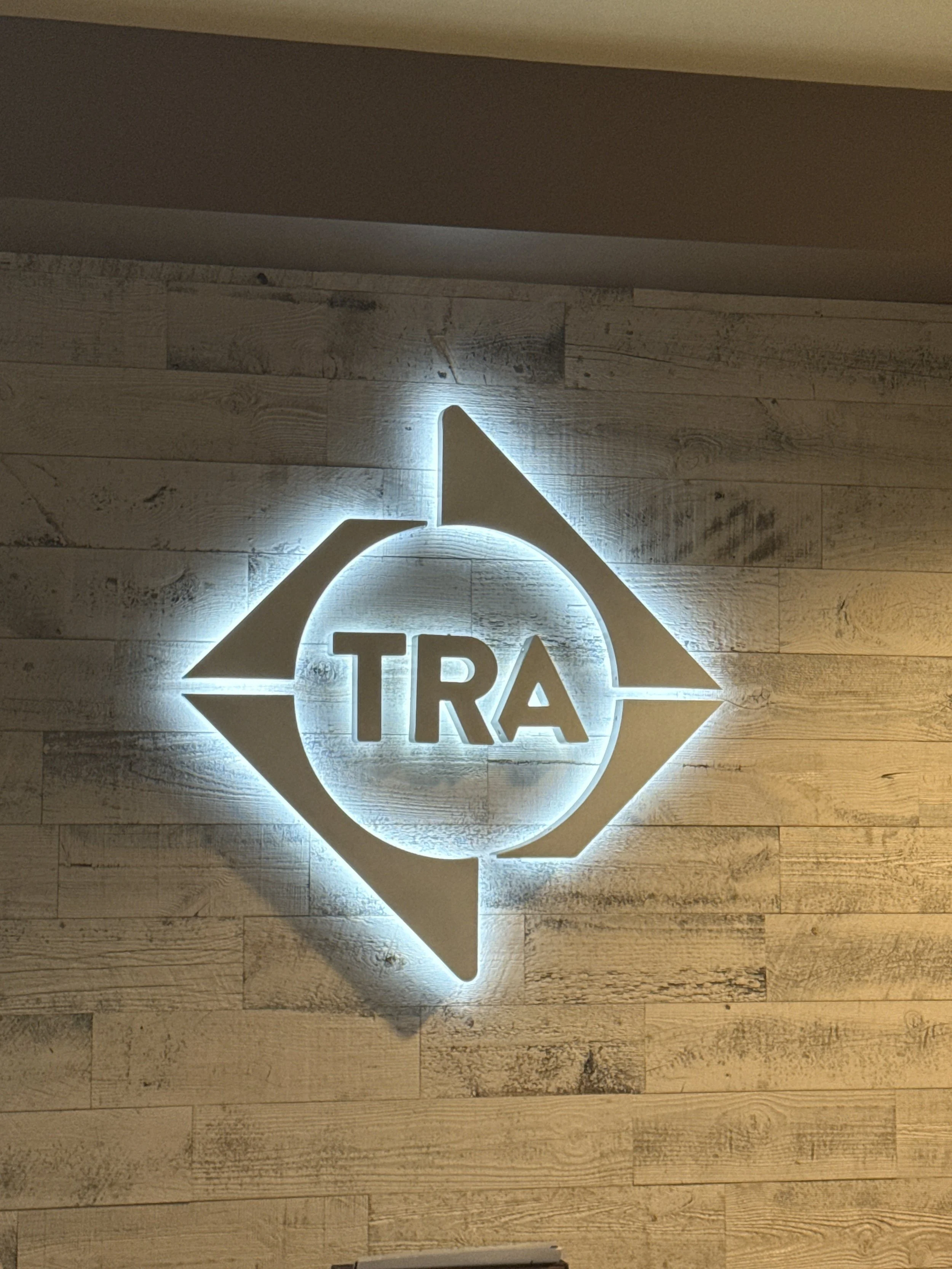 Halo-lit TRA diamond logo glowing on a horizontal wood-plank accent wall behind the front reception desk at TRA Medical Imaging's Gig Harbor office, fabricated and installed by Plumb Signs