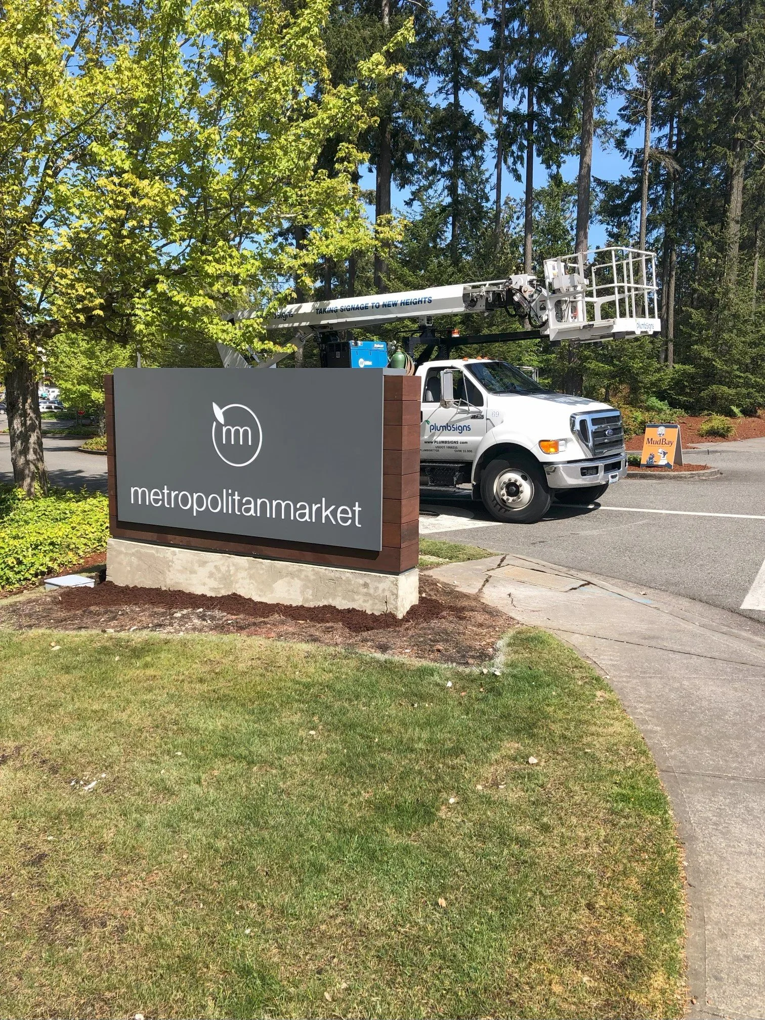 Plumb Signs install crew setting the double-faced Ipe-wood-skinned aluminum push-through monument sign onto its 10-foot finished concrete base at the Metropolitan Market parking lot, 5010 Point Fosdick Dr, Gig Harbor — routed-out copy backed with white acrylic and white LEDs