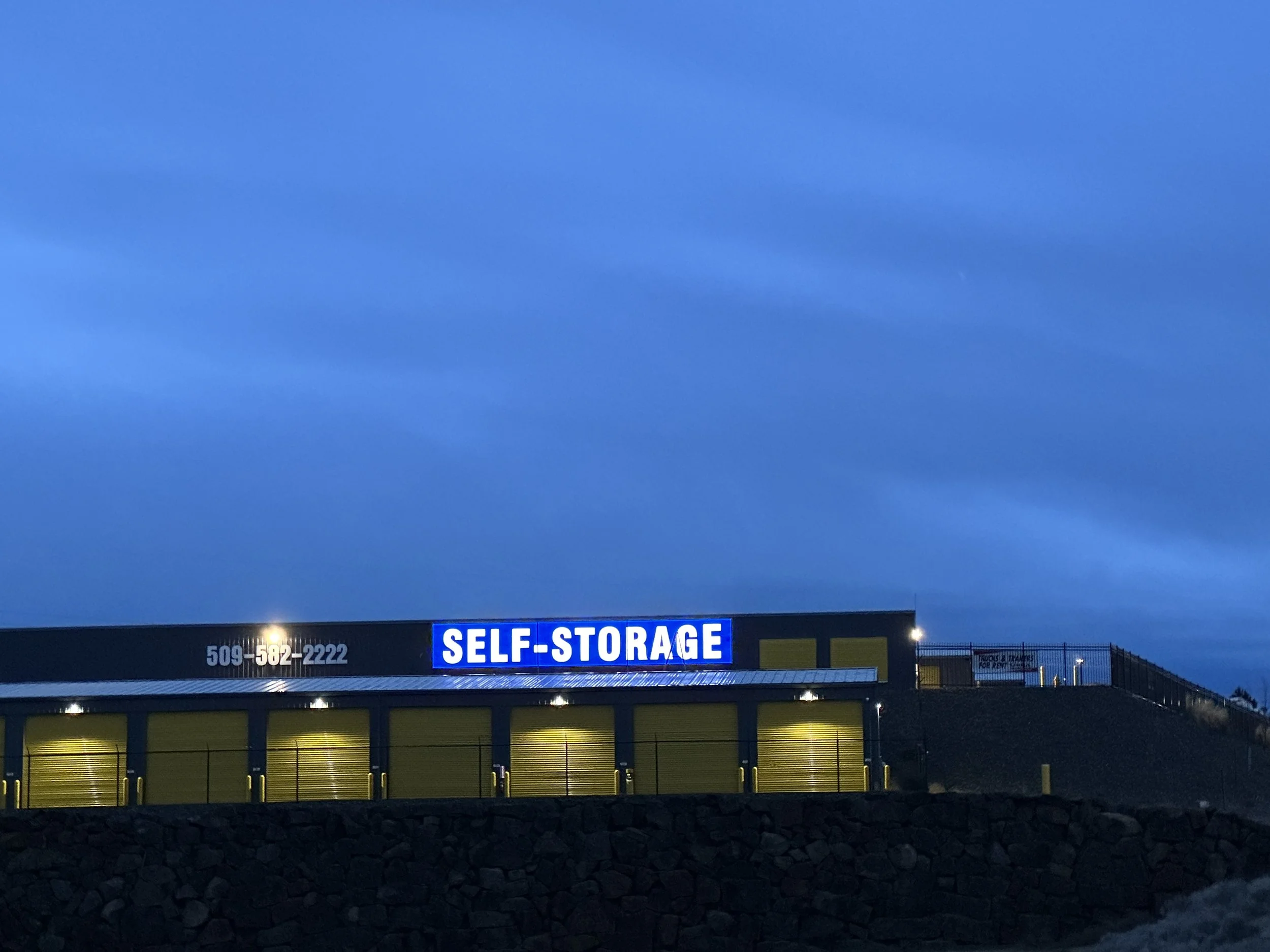 West Coast Self-Storage Kennewick property at 9501 W 10th Avenue — new 7-by-46-foot single-face flex-face wall cabinet illuminated at night alongside the adjacent phone-number cabinet, fabricated and installed by Plumb Signs