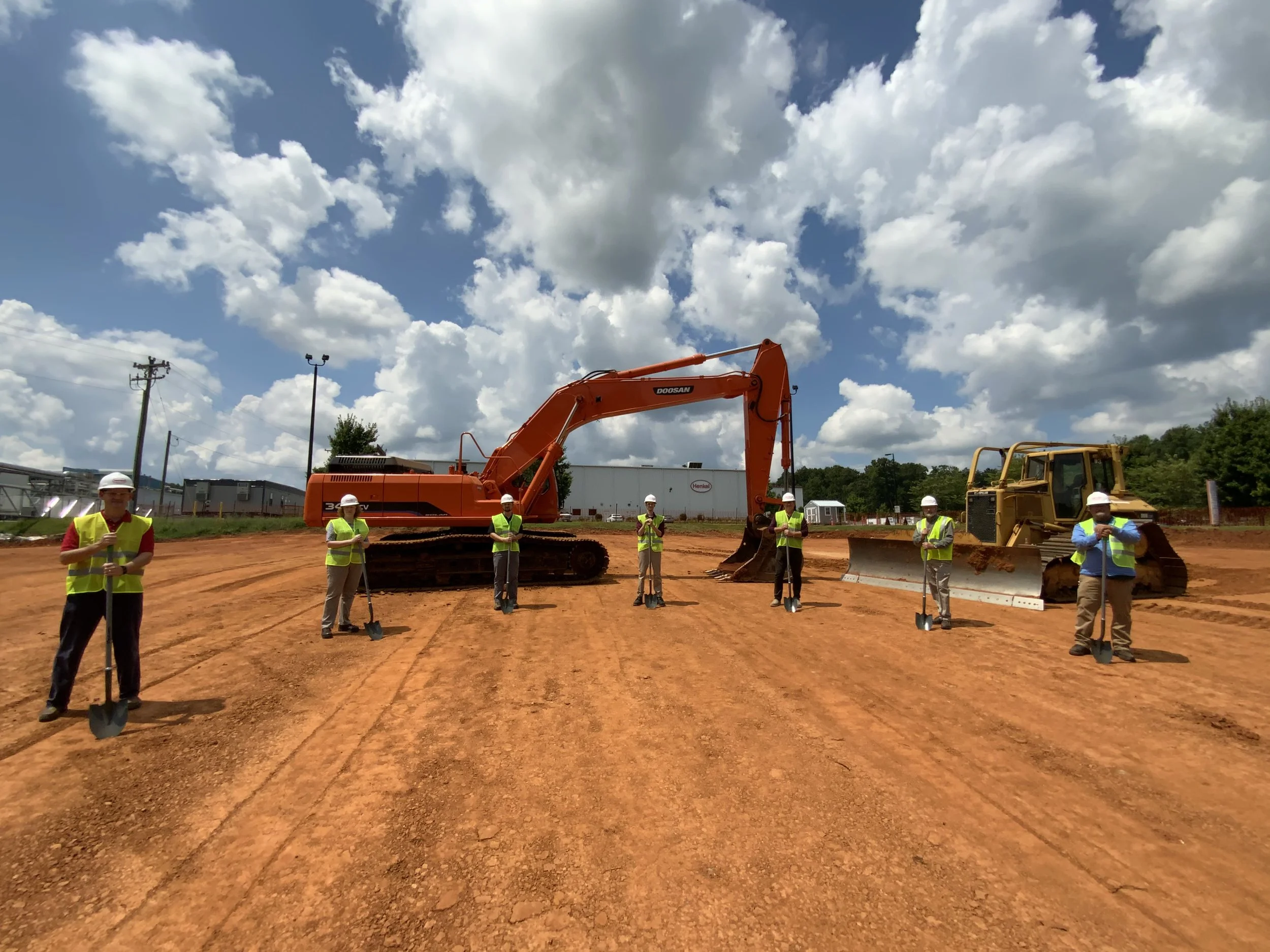 Seven people wearing hardhats and high-visibility vests hold shovels beside construction equipment